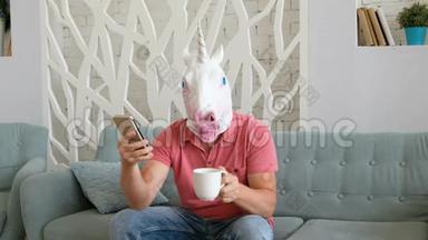 奇怪的搞笑视频：戴着独角兽面具的男人坐在沙发上拿着智能手机喝咖啡